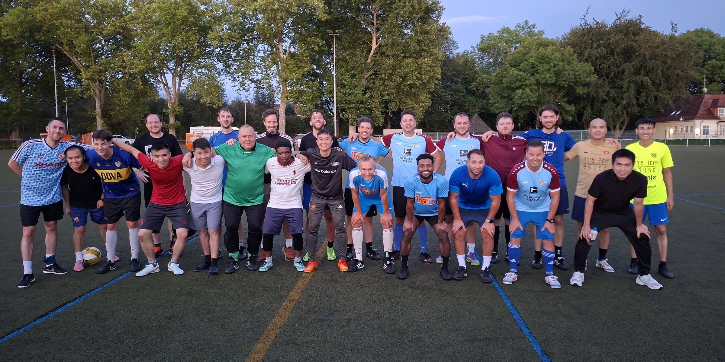 Gruppenfoto auf dem Kunstrasen am Abend: Spieler aus mehreren Teams der Liga in bunt gemischten Trikots, Arm in Arm nebeneinander.